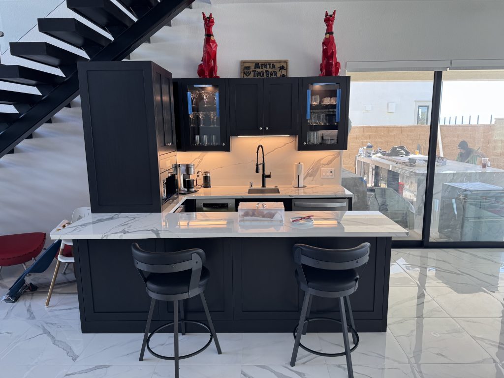 Custom under-stairs home bar in a Century City condo featuring matte black shaker cabinets, book-matched marble backsplash, and a modern black floating staircase with integrated glass shelving.